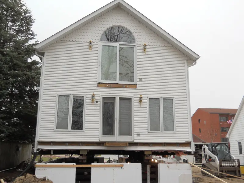 Two-story house lifted on steel beams and wood cribbing while a new foundation goes in underneath